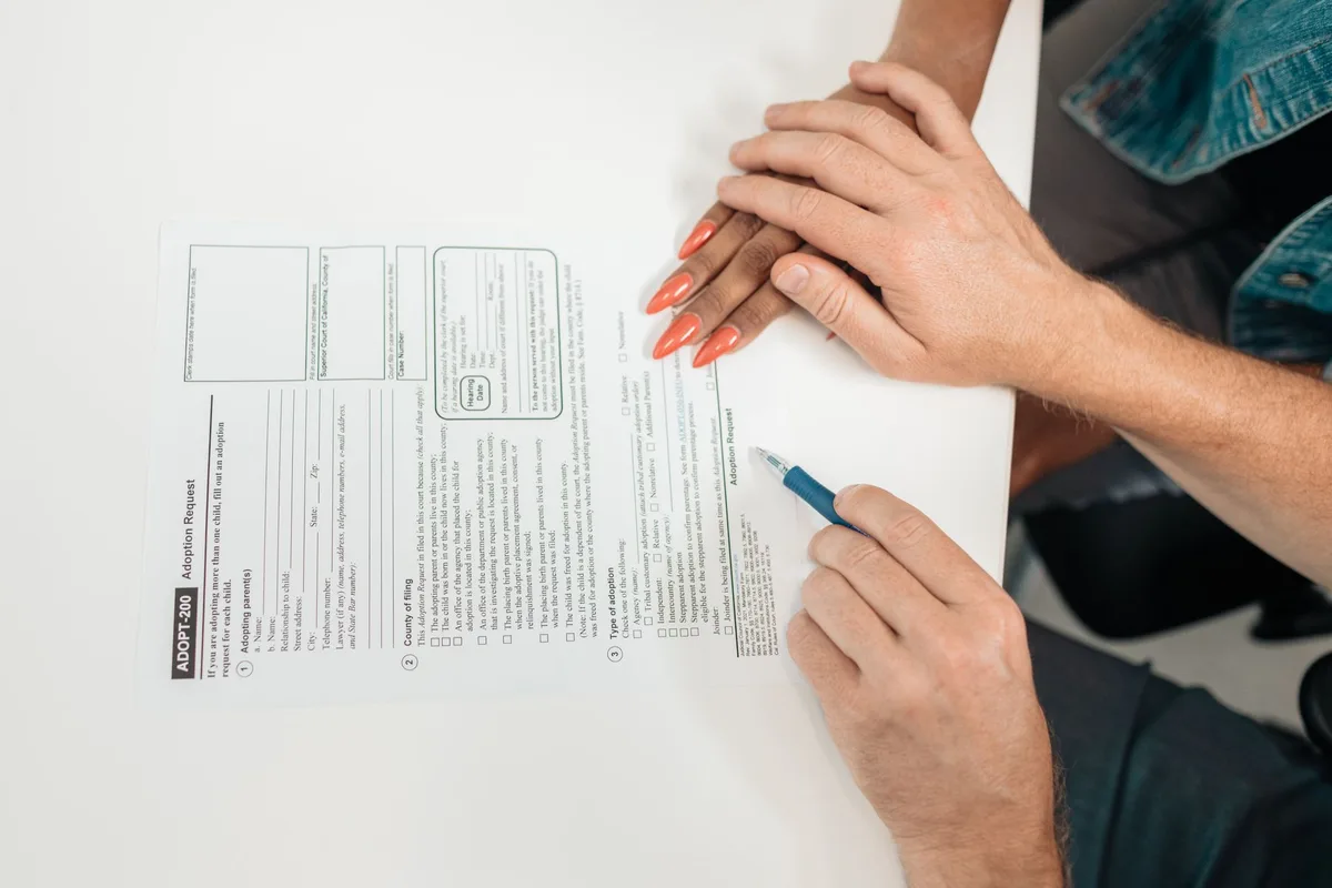 Hands filling a government application form on a wooden desk, paperwork and a closed pen nearby, no logos