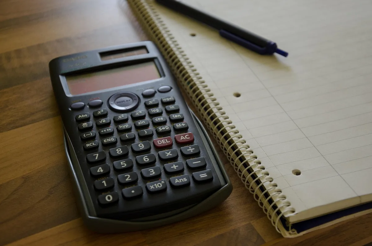 Close-up of hands holding a calculator and a budget sheet next to a small stack of documents, background softly blurred; no currency focus