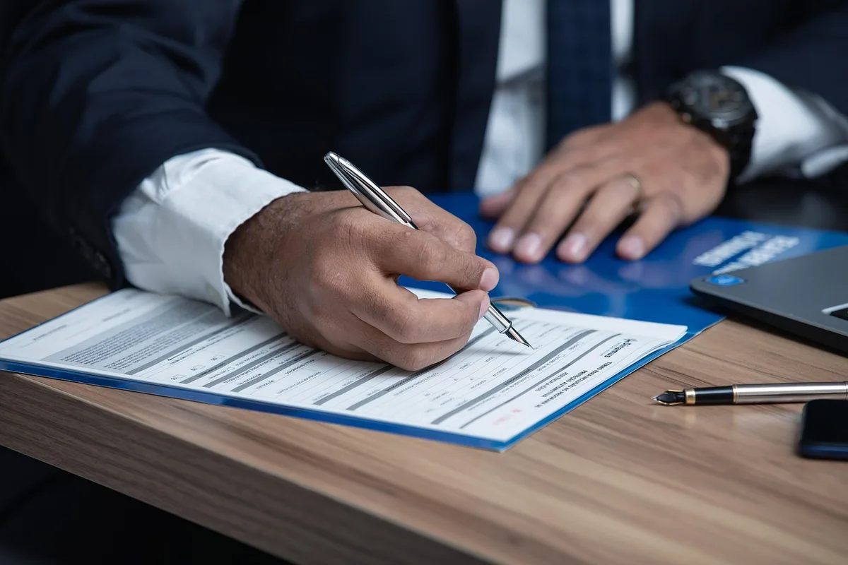 Hands filling a legal form on a wooden desk with a pen, a closed laptop and a blurred receipts stack in the background