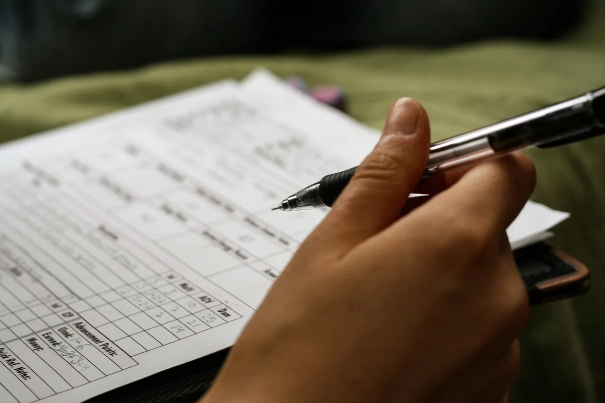 A well-lit desk with hands filling a printed application form, a pen and a closed notebook nearby, paperwork neatly arranged