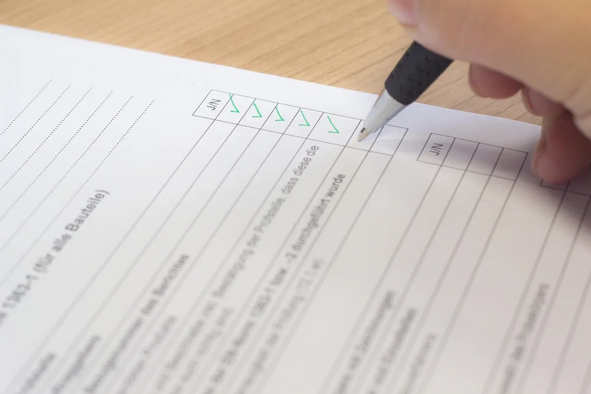 a desk with a printed certification list, a pen, and an anonymous pair of hands pointing at one line